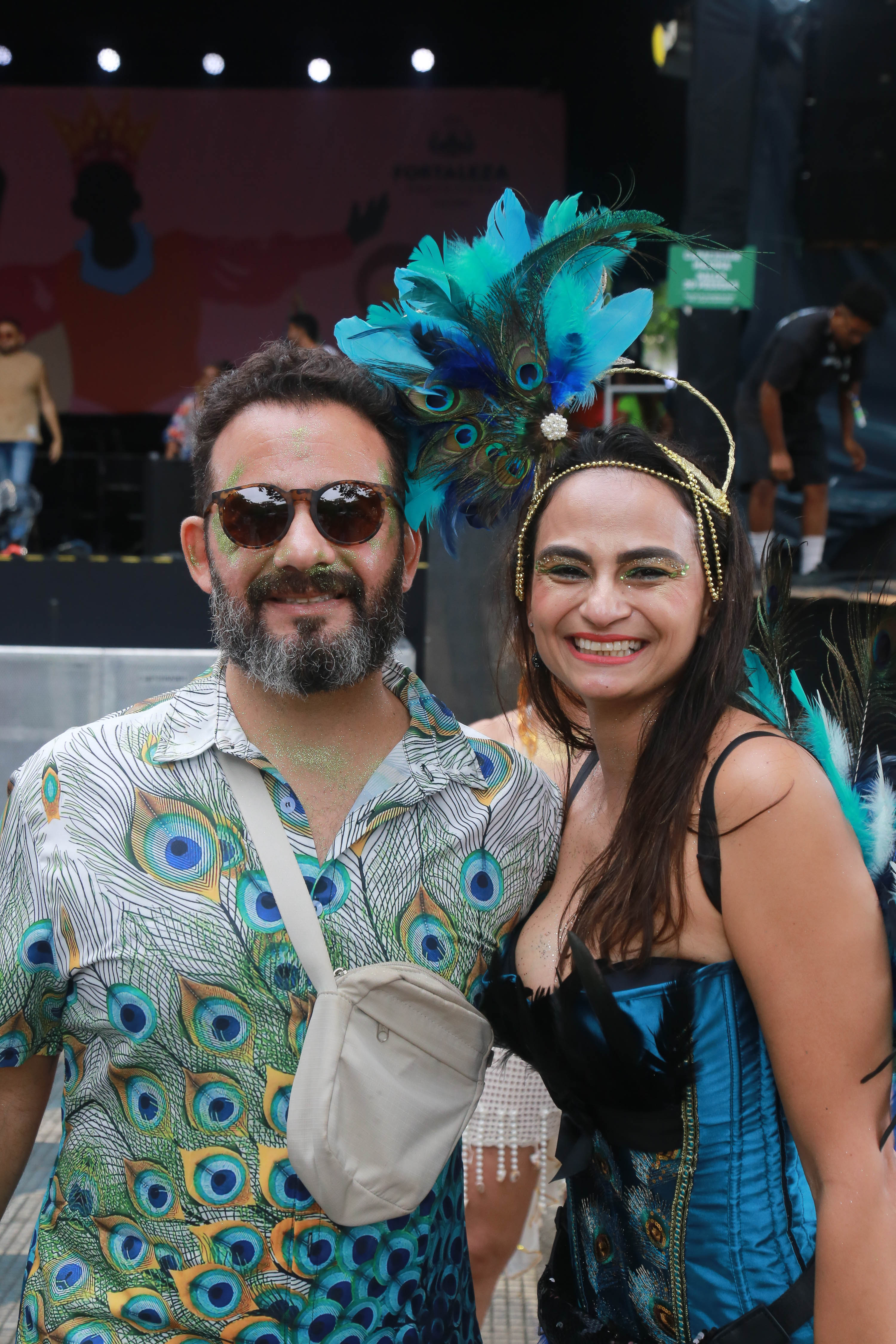 sara e joão paulo posam para a foto, ele está de camisa azul estampada e ela tem um arranjo na cabeça em forma de pavão